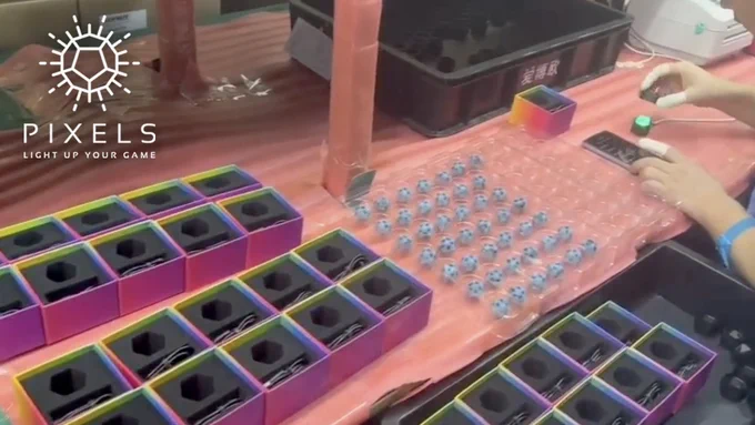 Video still: a worker at a table at a factory preparing dice for shipping. A tray of dice and open boxes are nearby while they program one with a mobile phone app. 