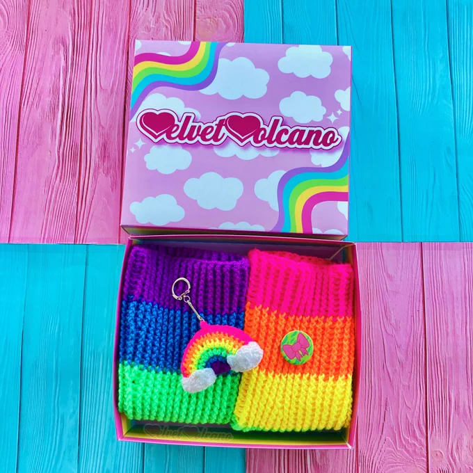 A pastel rainbow striped box with neon rainbow striped crochet leg warmers, a neon rainbow crochet 3D keyring with clouds at the ends and a green zebra print button badge with a pink bow on it.