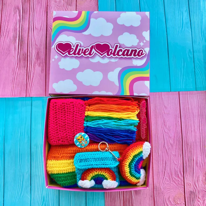 A pastel rainbow striped box with 3 crochet rainbow themed pieces including a rainbow striped scarf with rainbow tassels, a 3D stuffed rainbow and cloud keyring and a rainbow striped bum bag with a turquoise pocket on the front that features a rainbow and cloud motif and a turquoise adjustable strap. There is also a small turquoise button badge with a yellow kitty on it that’s wearing a rainbow and cloud themed top. The lid of the box is baby pink with white cloud print and wiggly pastel rainbows in the upper left and lower right corners, the pink & white VelvetVolcano logo is in the centre

