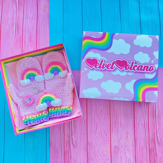 Baby Pink crochet mittens with pastel rainbow and cloud motif and pastel rainbow striped cuffs, a pastel rainbow with clouds at the ends crochet keyring and a pink badge with a cute pastel rainbow and cloud patterned teddy bear design in a pastel rainbow striped box with baby pink tissue paper inside. The lid of the box is baby pink with white cloud print and wiggly pastel rainbows in the upper left and lower right corners, the pink & white VelvetVolcano logo is in the centre