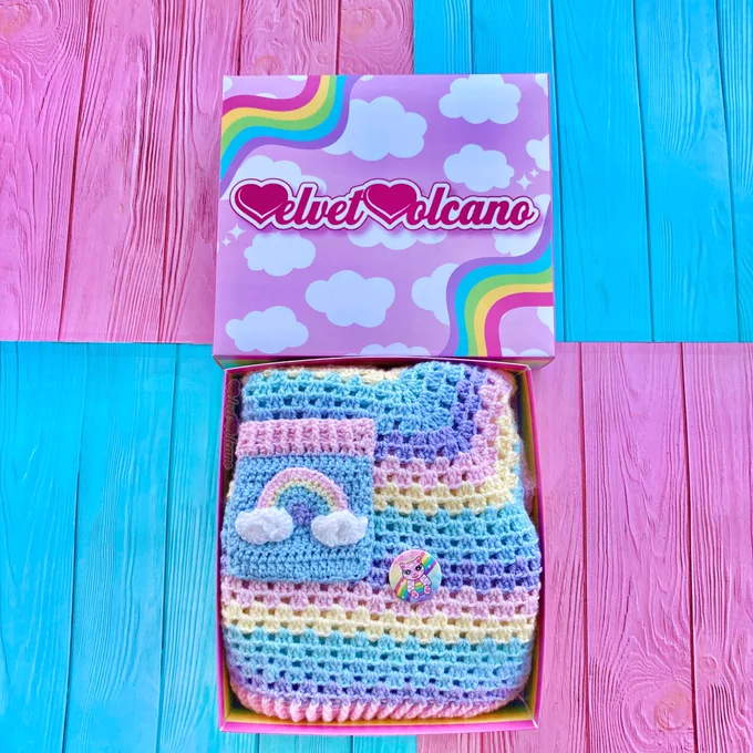 A pastel rainbow striped box with a crochet pastel rainbow striped sweater with a chest pocket that features a pastel rainbow with clouds at the ends. There is also a small pastel rainbow cloud kitty button badge. The lid of the box is baby pink with white cloud print and wiggly pastel rainbows in the upper left and lower right corners, the pink & white VelvetVolcano logo is in the centre.
