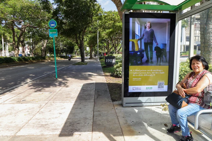 Photo of a digital billboard at a bus stop where a woman sits. The billboard features a photograph of Virginia Jacko with her guide dog Eva, the text "Who is Miami" and the quote "A blind person can do anything a sighted person does, but they might do it a little differently”.