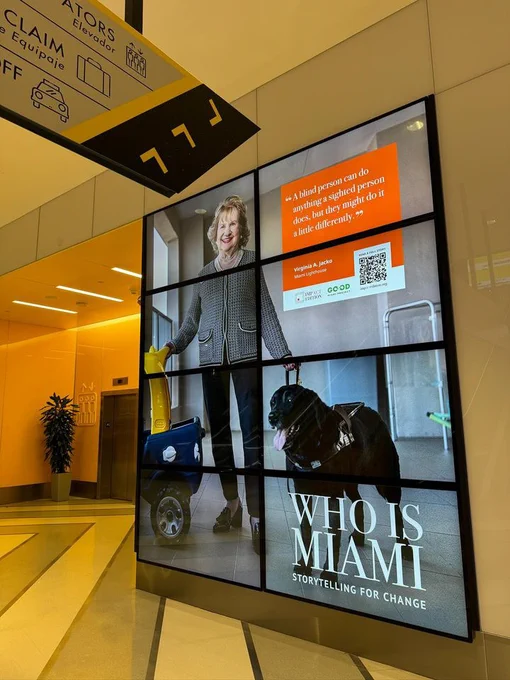 Photo of a digital billboard featuring at a train station, featuring a photograph of Virginia Jacko with her guide dog Eva, with the text "Who is Miami" and the quote "A blind person can do anything a sighted person does, but they might do it a little differently”.