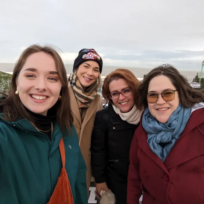 This is a selfie of Ruth, Claire, Vicky and Katie from Polyphony Arts. They are outside in front of the sea, all wearing winter coats and warm accessories.
