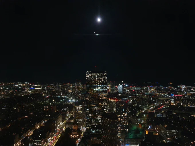 The view of Boston from the top of the Prudential centre. Lots of buildings and lights. The sky is dark; the moon is out.