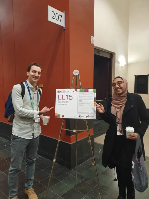 A man and woman standing on either end of a sign with "EL15 Chiral Materials - New Structures and Properties" written on it outside of a room with "207" written on top of the door and as a sign just outside. 