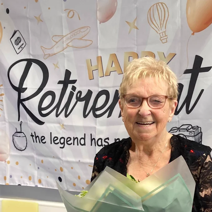 Norma stands in front of a sign that says 'Happy Retirement', she is holding flowers and smiling. 