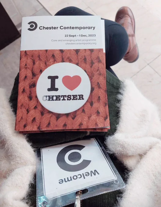 A photo looking down at a sitting person's knee. We can see a fluffy white cardigan, a lanyard with the word Welcome and the Chester Contemporary logo and a small pile of booklets about Chester Contemporary.

Photo: © Estelle Woolley.