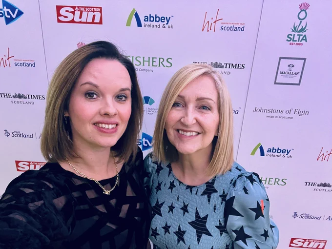 Anna and Lesley stand  in front of a display stand for the event. Anna has short brown hair to her shoulders and is wearing a black patterned dress. Lesley stands to her right with a blue dress covered in black stars and shoulder length blonde hair.