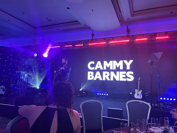 Cammy Barnes performing on stage - he stands at the mic playing his guitar with his name displayed behind him