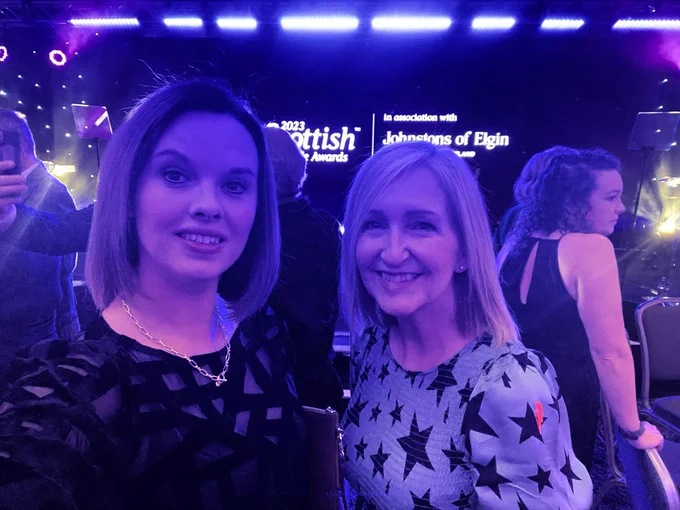 Anna and Lesley stand  in front of the stage for the event. Anna has short brown hair to her shoulders and is wearing a black patterned dress. Lesley stands to her right with a blue dress covered in black stars and shoulder length blonde hair.