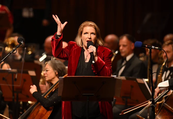 Clare Teal singing into a microphone. Her right hand is raised in a gesture and she is wearing a long red velvet jacket. Members of the BBC Concert Orchestra Orchestra are seated behind her.