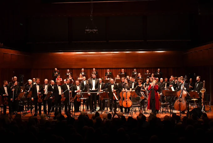 The BBC Singers, BBC Concert Orchestra, conductor Owain Park and presenter Clare Teal stand on the Milton Court Concert Hall stage, facing an applauding audience. They are smiling.