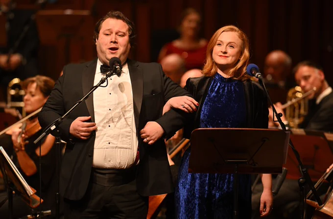 Two members of the BBC Singers, tenor Peter Davoren and alto Nancy Cole, stand arm in arm whilst singing.
