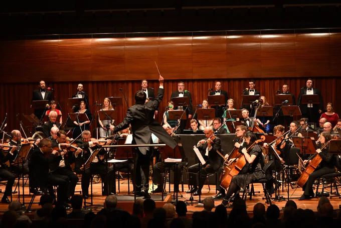 The BBC Concert Orchestra and the BBC Singers performing on the Milton Court Concert Hall stage, conducted by Owain Park who has his arm raised, holding a baton, his back to the camera, coattails sweeping out to the side as he gestures.
