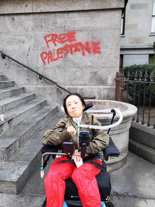A photo of an Asian American disabled woman in a wheelchair with a tracheostomy attached to a ventilator. She is wearing an olive bomber jacket and red pants. Behind her is a building FREE PALESTINE written in red paint