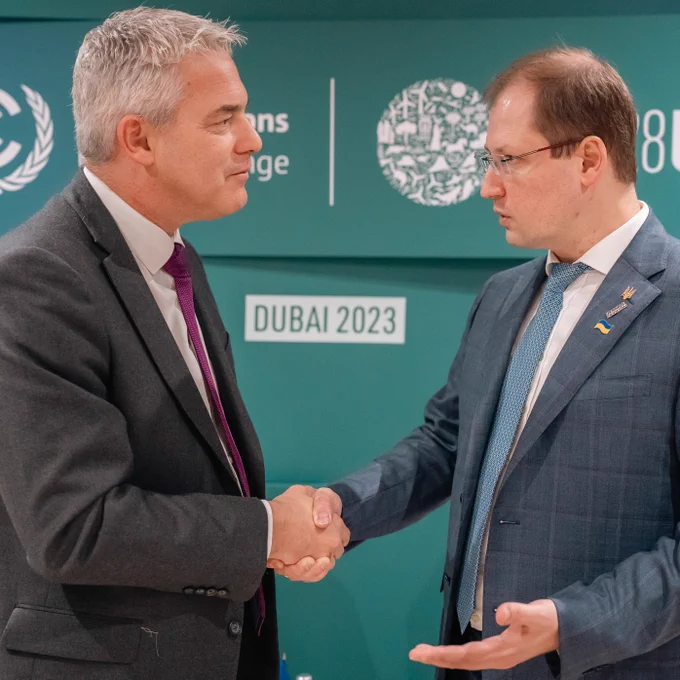 The UK's Environment Minister Steve Barclay and the Ukrainian Environment Secretary Ruslan Strilets shake hands in front of a green wall with the COP28 logos partially visible behind them. Both men wear suits and ties. They are standing in front of one another and are looking directly at each other. 