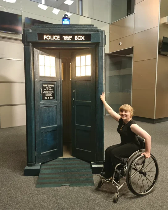 Ruth in her wheelchair next to the big blue Tardis with a ramp coming out of it. Ruth is wearing a black jumpsuit & her hair is tied in a chignon.