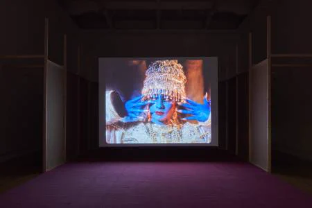 An installation of a large film screen and pink carpet. The screen shows an actor painted blue and dressed in a silver beaded garment. 