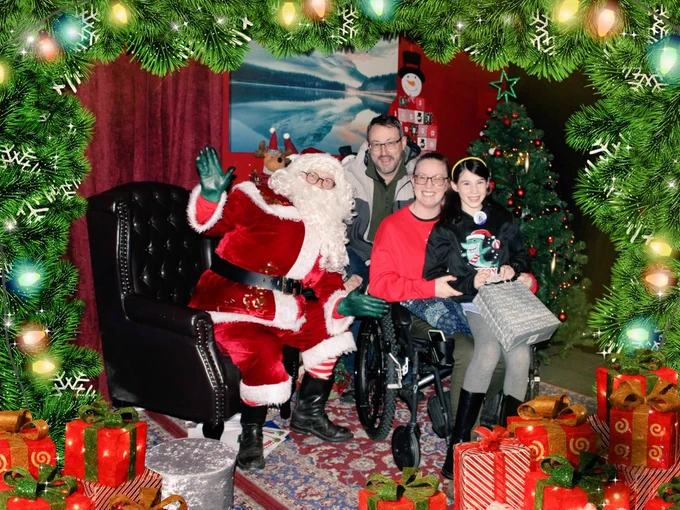 Family photo with Santa in his grotto, showing mum, dad and girly. Girly sits on mum's lap and is wearing a sharky Christmas jumper. All are smiling and Santa is waving.