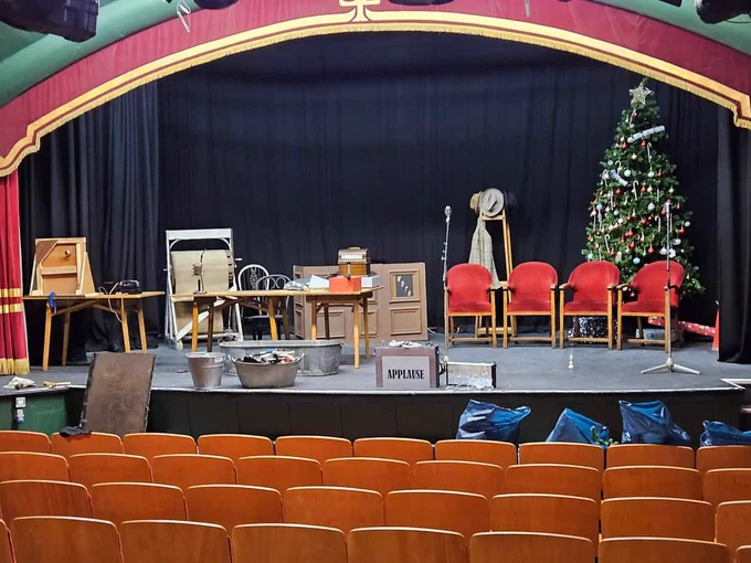 A photograph of the stage at the Little Theatre Gateshead set up for the radio play. There is a fully decorated Christmas tree, chairs, a piano and an assortment of items to make noises during the show.