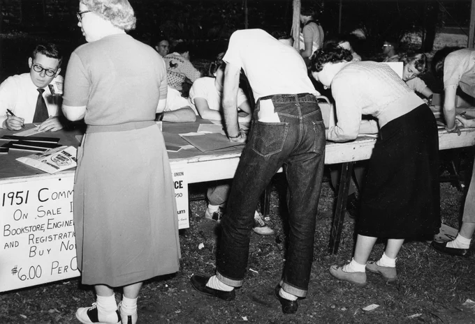 1951 Commodore Yearbook sales, four students stand at a row of tables to sign up for yearbooks. About five students sit at the tables to assist the standing students. Poster at the bottom left of the photo states that the yearbook is available for $6. Black and White photograph