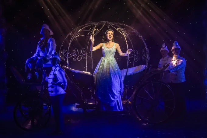 Cinderella (Clair Gleave) in a blue sparkly dress sings from a golden carriage surrounded by 3 ensemble members in white and golden outfits with Buttons (James Merry) sitting at the front of the carriage with a coach drivers hat. 
