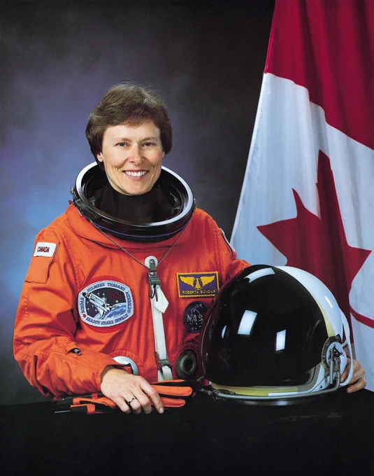 Dr. Roberta Bondar in an orange flight suit with a Canada flag in the background. 