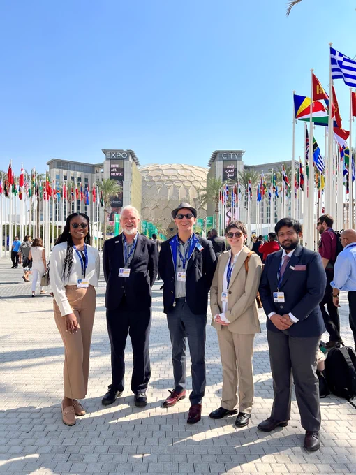 Five people standing in Dubai, a pedestrian area behind them is flanked by dozens of national flags. In the background is a beige builidng that looks like a honeycombed sphere sat between two tower blocks which read 'EXPO CITY'