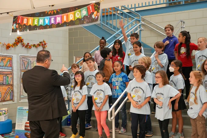 Secretary Cardona speaks with a group of elementary school students standing under a banner that reads "Bienvenidos!"