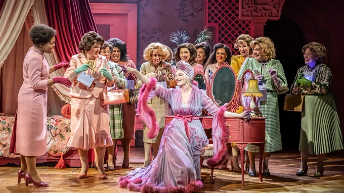 A woman in a purple dressing gown and head scar with pink feather lining sits on a stool at a dressing table. A large group of women in 1950s esque pastel clothes stand around her. Two take a selfie with her. There is a grand four poster bed behind them. The women hold small purple gift bags. 
