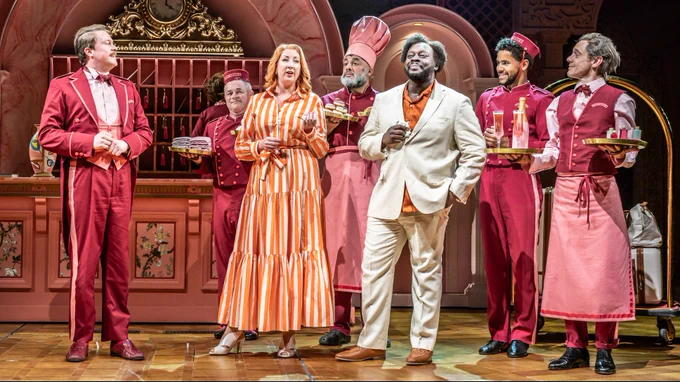 A woman in an orange and white striped dress and a man in and orange and white suit stand side by side. Behind them a group of people in pink hotel uniform for maids and bell boys etc stand in a line.  They are in a pink and gold stage set of a hotel reception.