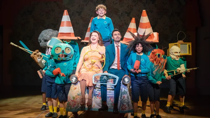 A woman in an orange tea dress sits next to a man in a blue suit with orange tie. She holds a steering wheel and he holds the front grate of a car to give the illusion of driving a car. A boy in a blue jumper and yellow shirt stands behind them. Other children in the same hat with traffic cones on their heads stand around them. 