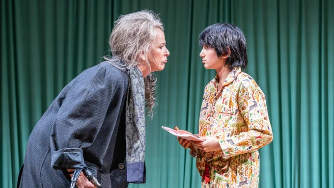 A boy in patterned pyjamas holds a piece of paper. A woman with grey hair makes a mocking sad face at him.