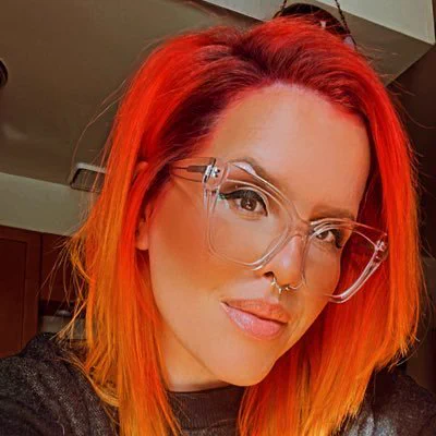 Anya Combs, woman with intensely red hair, large clear framed glasses, nose piercings, headshot appears to be indoors but not much of the background is visible