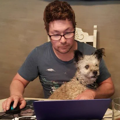 Ryan Ike, short haired fit guy wearing glasses with a dog on his lap as he sits focused on a laptop with his hand on the mouse