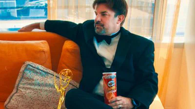 Playfully serious looking man, Mike Mika, dressed in a tuxedo with an important golden award trophy next to him (golden globe, maybe?) but also a can of spicy looking pringles chips in his hand, looking off to the side with his arm over the back of the couch