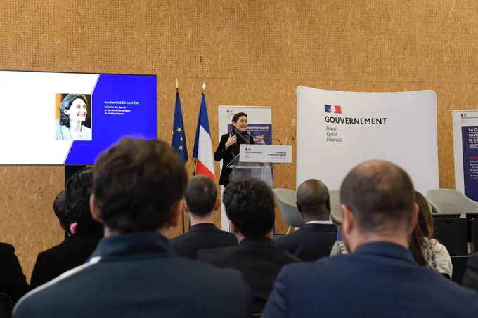Au premier plan, des personnes de dos regardent Amélie Oudéa-Castéra qui prononce son discours. 
Au second plan, sur la gauche de la ministre un écran diffuse un diaporama, au centre Amélie Oudéa-Castéra s’exprime derrière un pupitre, à droite un fond de scène blanc avec le logo Gouvernement