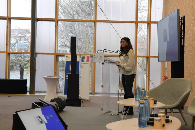 Sur l’estrade, au premier plan, un écran en bas à gauche, une table et un fauteuil sur la droite. 
En arrière, Prisca Thevenot s’exprime derrière un pupitre