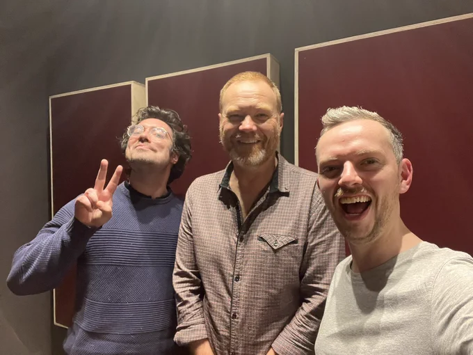 Photo in the studio of Sam giving the peace sign, next to Stephen and Dave Smiling.