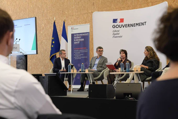 Au premier plan des personnes de dos écoutent les intervenants sur la scène. 
Au second plan, quatre intervenants échangent dans le cadre de la première table ronde dédiée au sujet « le service civique dans le sport »