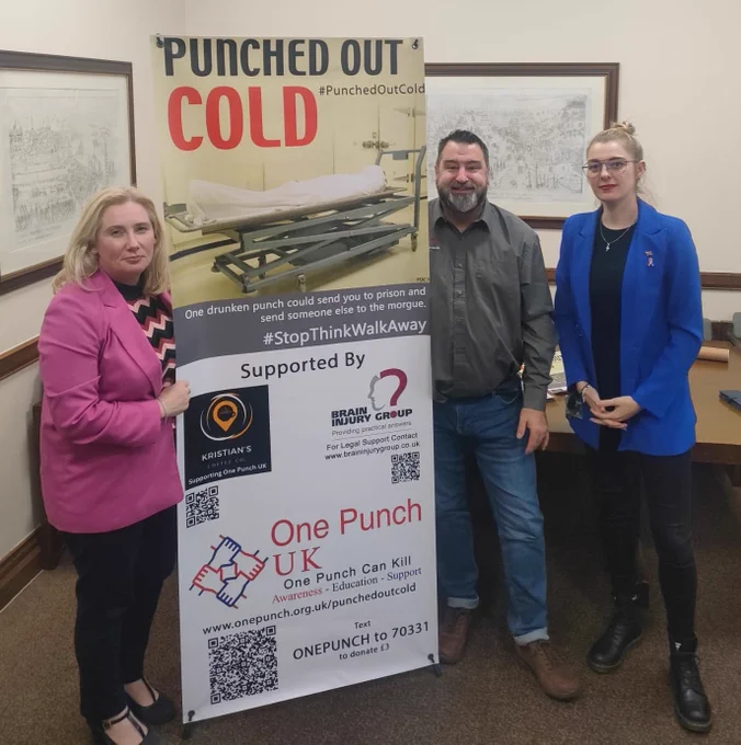 This picture shows Emma Lewell-Buck MP in with two people next to a banner for One Punch UK. On the banner is a picture of a covered body on a gurney with the caption "punched out cold" and the hashtag 'stop think walk away'. The One Punch UK charity logo is visible beneath. 