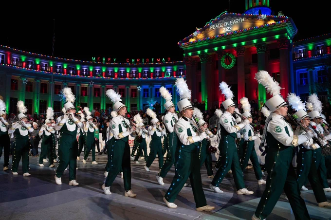 The Colorado State University marching band plays instruments while marching down the street. The building behind them is adorned with Christmas lights, including text that reads "Merry Christmas" and "Peace On Earth 2023."