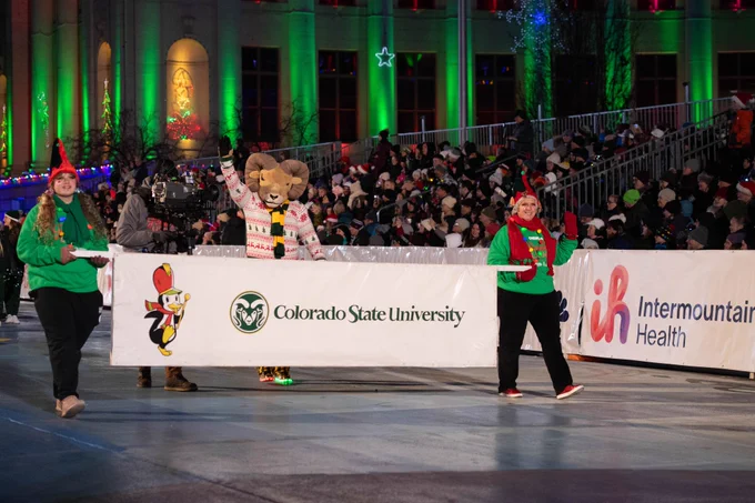 Two people carry a Colorado State University banner as part of the Parade Of Lights event. CAM The Ram joins the festivity, wearing a holiday-themed sweater and waving to the crowds. The two banner carriers are adorned in elf hats, adding a joyful touch to the scene.