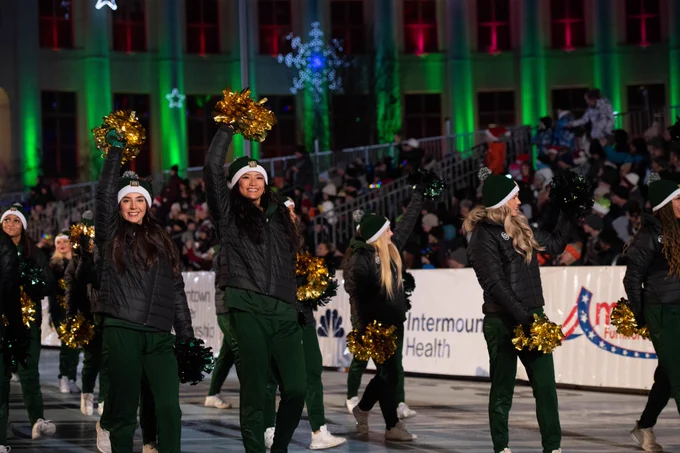 The CSU cheer squad adds festive cheer to the Parade of Lights, waving golden pompoms as they march down the street. The building behind them is aglow with Christmas lights.