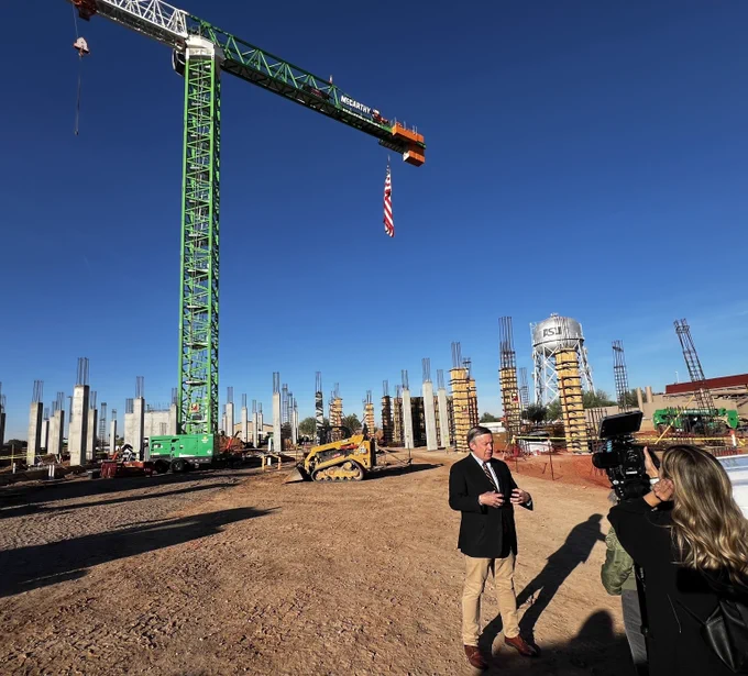 A person in business attire gives an on-camera interview in front of a construction crane at the site of a new campus engineering building.