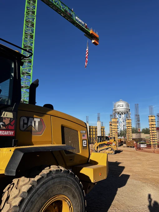 A bright yellow construction vehicle sits next to a large crane displaying the American flag in front of an ASU water tower at a construction site.
