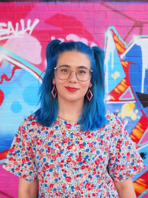 head shot photo of rowan - a white woman with glasses and blue hair in bunches, wearing bright pink earrings, and a blue and pink shift dress. she's stood in front of a blurry graffiti wall and is smiling softly at the camera.