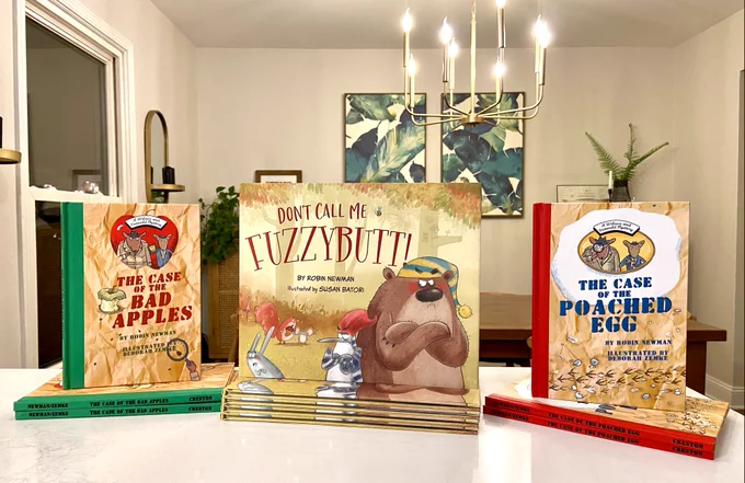 Photograph of 11 books for young readers stacked in several piles with three books face out. The book are THE CASE OF THE BAD APPLES, DON'T CALL ME FUZZYBUTT!, and THE CASE OF THE POACHED EGG. The books are on a white kitchen countertop. The wall beyond is light gray and is decorated with a gold sconce and two large paintings of green palms. A brass chandelier hangs from the ceiling.