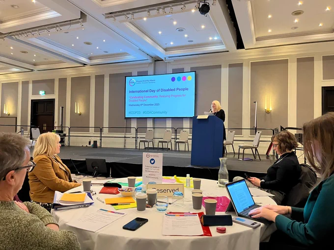 Image of GDA CEO Tressa stood at a blue podium, looking out at the room of 100  GDA members and addressing the room.